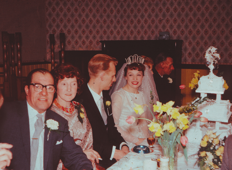 A bride and her groom sit at a table with cake and family in the 1960s